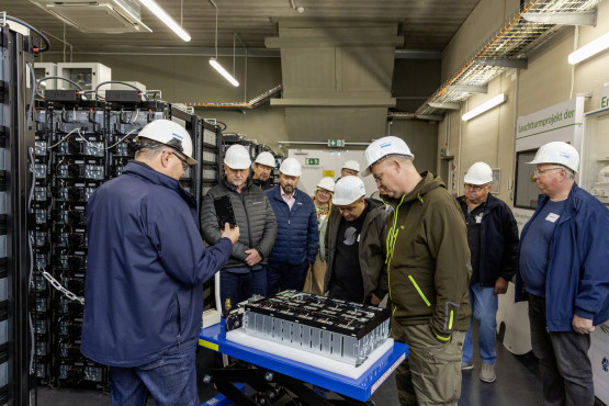 Batteriespeicherexperte Tobias Struck im Vordergrund steht an einem Batteriespeicher-Modell und erklärt den Mitgliedern des WEMAG-Kundenbeirats die Funktionsweise. Alle tragen weiße Helme.